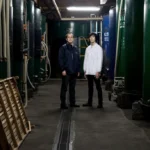 Two sake brewers from Kurosawa Brewery standing in industrial facility with large tanks.