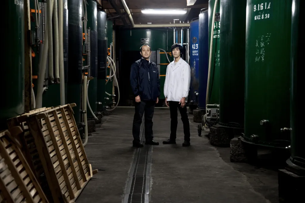 Two sake brewers from Kurosawa Brewery standing in industrial facility with large tanks.